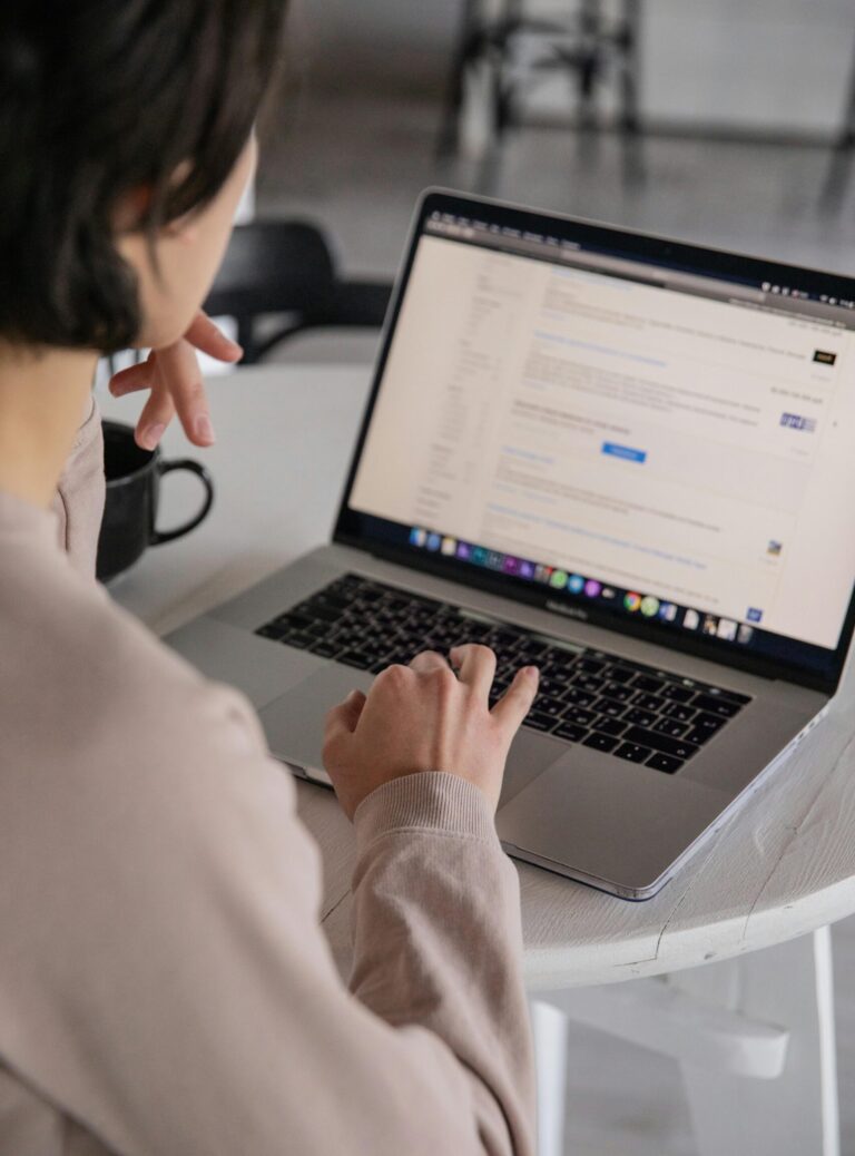 Back view of crop anonymous female remote employee typing on portable computer while watching website on screen and sitting at plastic table with cup of hot drink in apartment